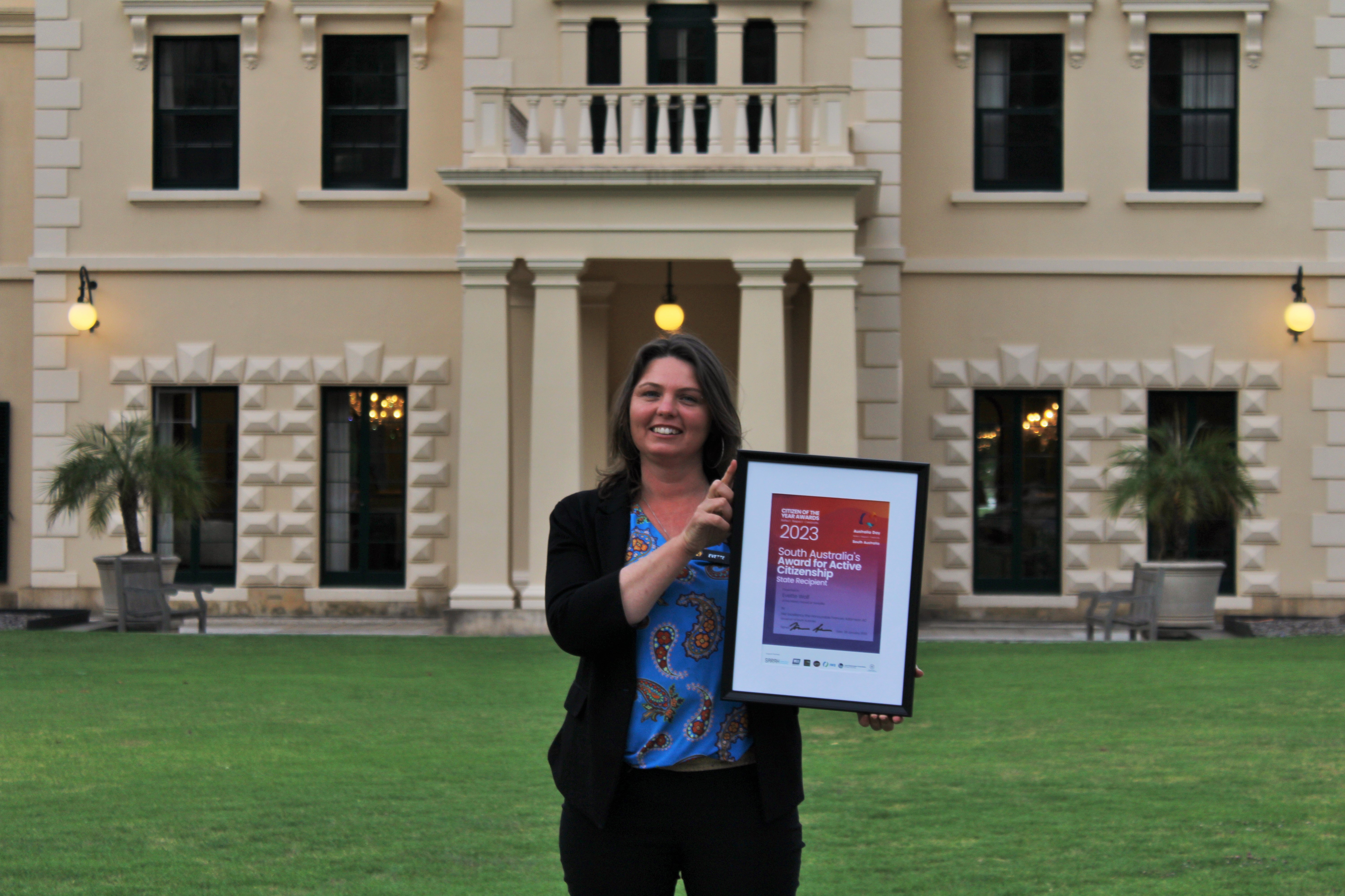 An image of Evette Wolf standing out the front of Government House, holding a certificate and smiling.