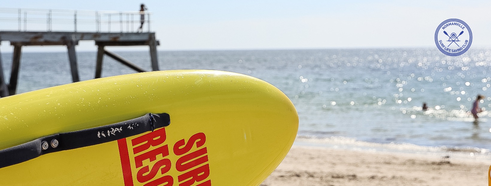 A photo of the Normanville Beach with a rescue board in the foreground and the surf club logo in the top corner