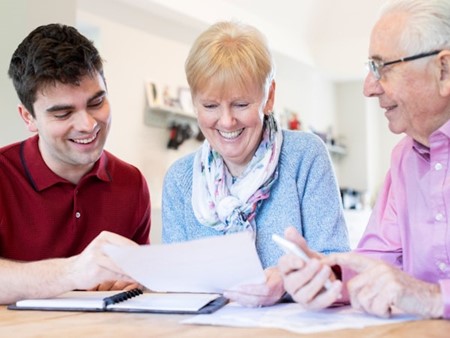 Two elderly people (a man and a women) are being assisted to complete some forms by a young man. They are all smiling.